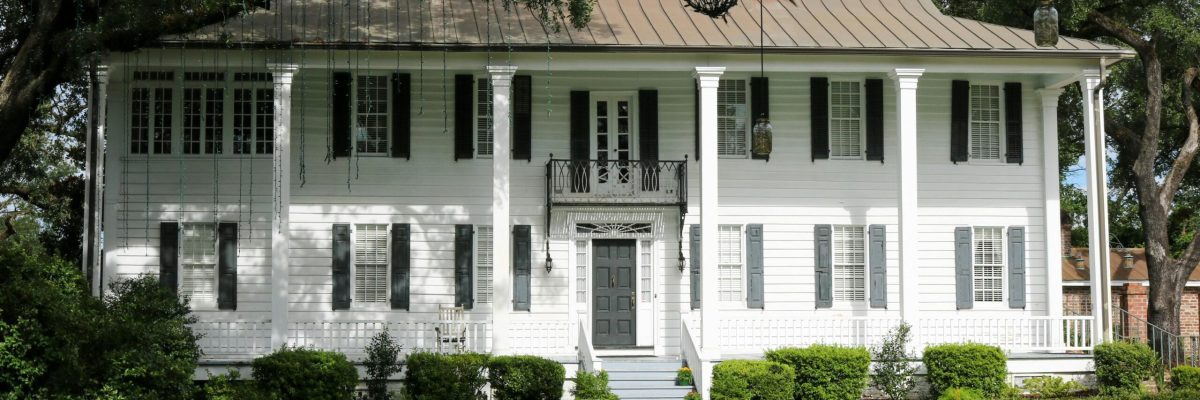 Elegant colonial house with lush lawn and classic facade surrounded by trees.